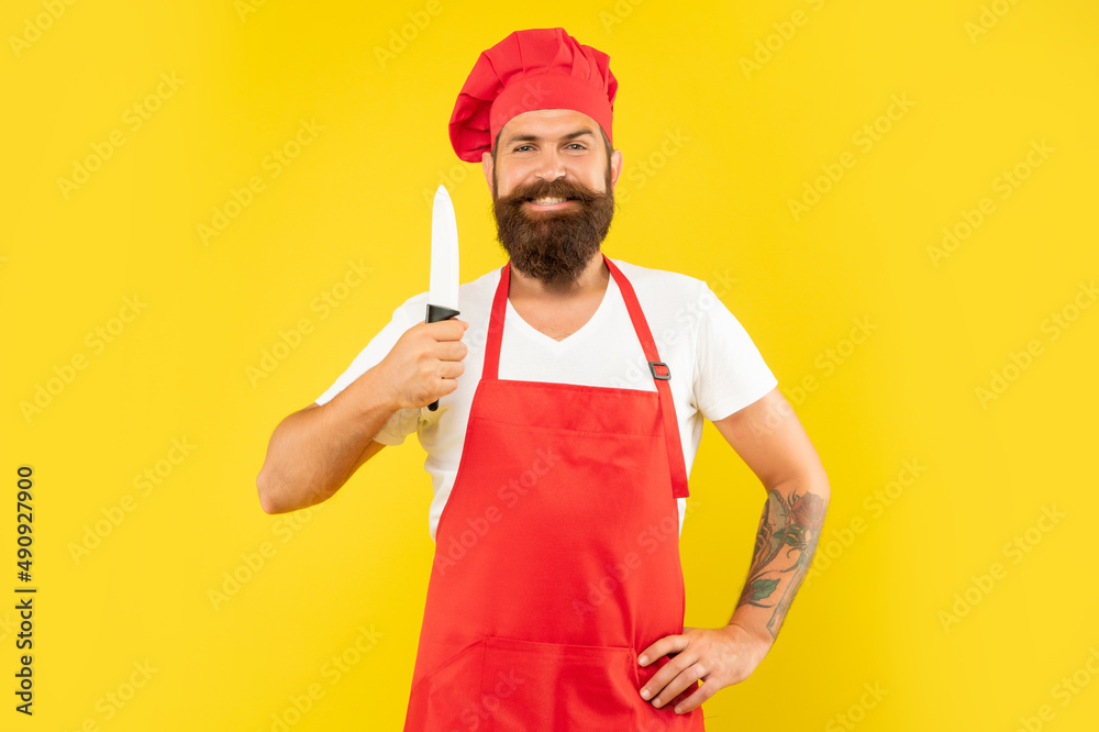 Happy man in red apron and toque holding chefs knife yellow background, chef