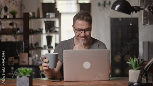 Bearded man working online with laptop computer at home sitting at desk. Home office, browsing internet. Portrait of mature age, middle age, mid adult man in 50s.