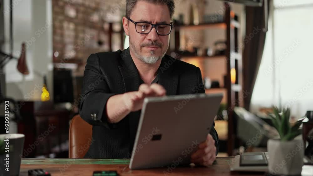 Older man sitting at desk in dark cosy room working on tablet computer in home office. Mature age, middle age, mid adult casual man in 50s, confident. Older man sitting at desk in dark cosy room working on tablet computer in home office. Mature age, middle age, mid adult casual man in 50s, confident.