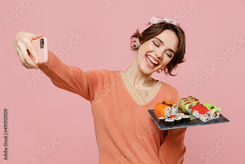 Young fun happy woman wear sweater hold in hand makizushi sushi roll served on black plate traditional japanese food doing selfie shot on mobile cell phone isolated on plain pastel pink background
