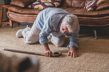 Elder senior man lying on floor after falling down with wooden walking stick beside couch on rug in living room at home. Old man suffering with pain and struggling to get up after falling down 