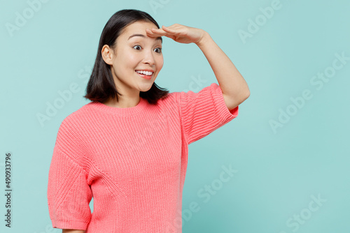 Young smiling happy woman of Asian ethnicity 20s wearing pink sweater hold hand at forehead look far away distance isolated on pastel plain light blue color background studio People lifestyle concept
