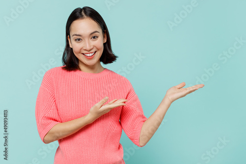 Young smiling happy woman of Asian ethnicity 20s wearing pink sweater hands arms finger aside on workspace area mock up isolated on pastel plain light blue background studio. People lifestyle concept.