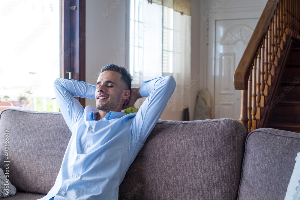 Satisfied young man relaxing while sitting on couch with hands behind head at home. Handsome smiling male sitting comfortably on sofa in living room of modern house, feeling peace of mind Satisfied young man relaxing while sitting on couch with hands behind head at home. Handsome smiling male sitting comfortably on sofa in living room of modern house, feeling peace of mind
