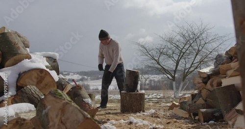 a middle-aged European man, dirtily dressed, straightens a log, takes an ax, looks into the camera and splits firewood in a village against the backdrop of winter mountains. overall plan