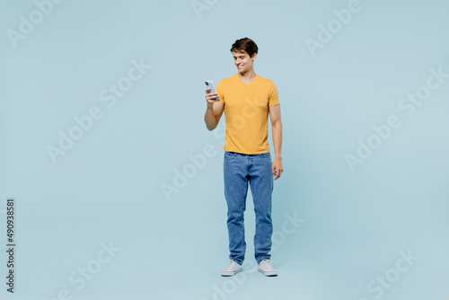 Full body young man 20s wearing yellow t-shirt hold in hand use mobile cell phone browsing surfing internet isolated on plain pastel light blue background studio portrait. People lifestyle concept