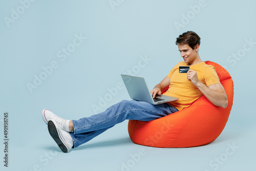 Full body young man in yellow t-shirt sit in bag chair using laptop pc computer hold credit bank card doing online shopping order delivery booking tour isolated on plain pastel light blue background