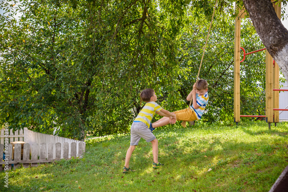 Two adorable happy little boys is having fun on a rope swing which he has found while having rest outside city. Active leisure time with children