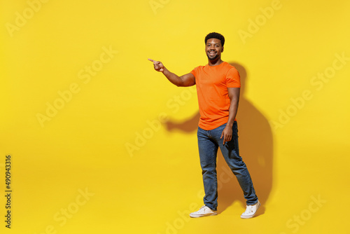 Full body side view smiling young man of African American ethnicity 20s wear orange t-shirt walking go point index finger aside on workspace area isolated on plain yellow background studio portrait