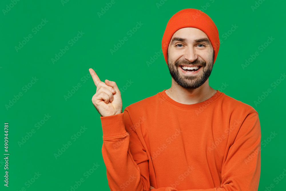 Young smiling happy fun caucasian man 20s wear orange sweatshirt hat point index finger aside on workspace area mock up isolated on plain green background studio portrait. People lifestyle concept Young smiling happy fun caucasian man 20s wear orange sweatshirt hat point index finger aside on workspace area mock up isolated on plain green background studio portrait. People lifestyle concept