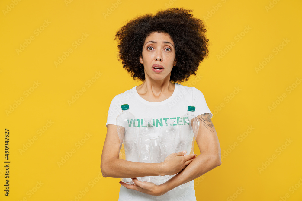 Young shocked woman of African American ethnicity wears white volunteer t-shirt hold water plastic pet bottles isolated on plain yellow background. Voluntary free work assistance help grace concept.
