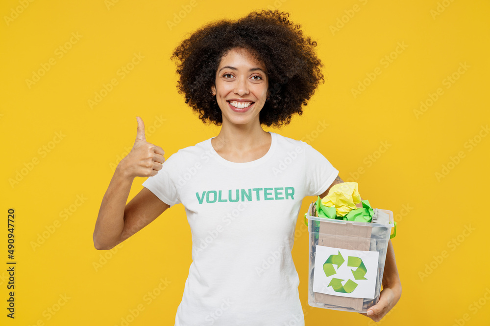 Young woman of African American ethnicity in white volunteer t-shirt hold boxes for waste sorting show thumb up isolated on plain yellow background. Voluntary free work assistance help grace concept.