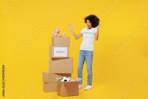 Full body surprised young woman of African American ethnicity wears white volunteer t-shirt near boxes with presents isolated on plain yellow background. Voluntary free work assistance help concept. photo
