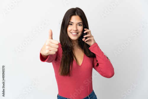 Teenager Brazilian girl isolated on white background keeping a conversation with the mobile while doing thumbs up