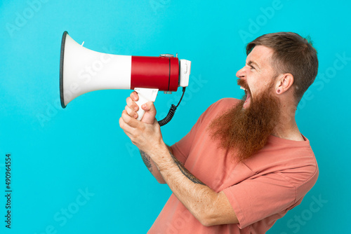 Young reddish caucasian man isolated on blue background shouting through a megaphone to announce something in lateral position © luismolinero