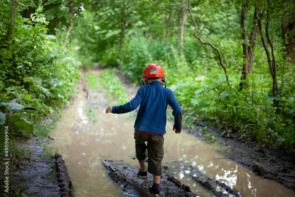 Child in nature. Boy plays near puddle. Preschooler travels in wilderness. 6-year-old boy stands on muddy road in woods.