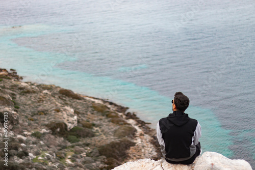 Back view of sitting young man on rocks. He is raising both hands and doing peace sign with fingers. Sea background.