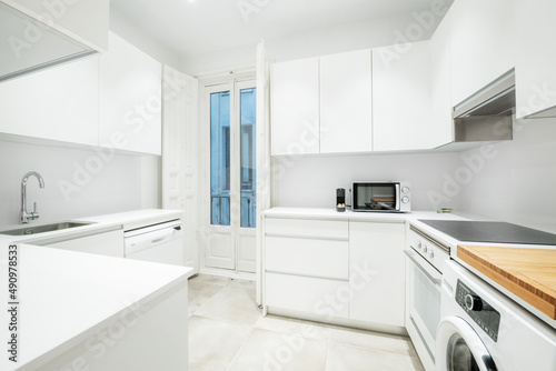 Nice kitchen of a short-term rental apartment with white furniture and appliances  with a balcony with vintage wooden doors and a ceramic hob