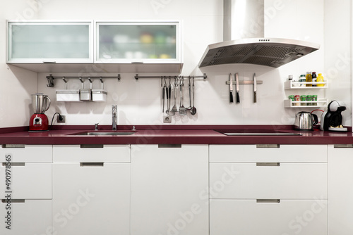 Front image of a kitchen with white lacquered furniture, a red countertop and many accessories on the walls and on the countertop