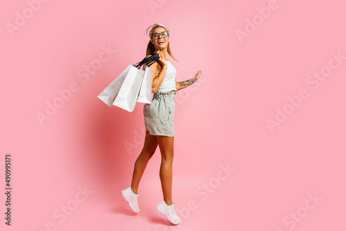  Excited blond woman holding white shopping bags and posinh on pink background. Spring season. Full lenght.