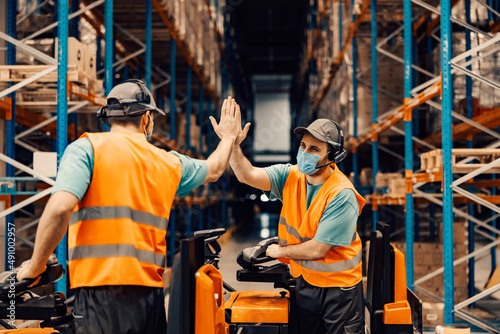 Fotografie, Obraz Two delivery service workers with voice picking headset on forklifts giving high five for collaboration during corona