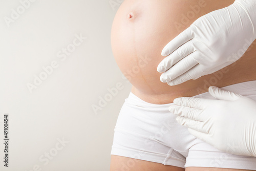 Doctor hands in white rubber protective gloves touching young pregnant woman big belly skin on light gray background. Examining caesarean scar before second baby birth. Closeup.