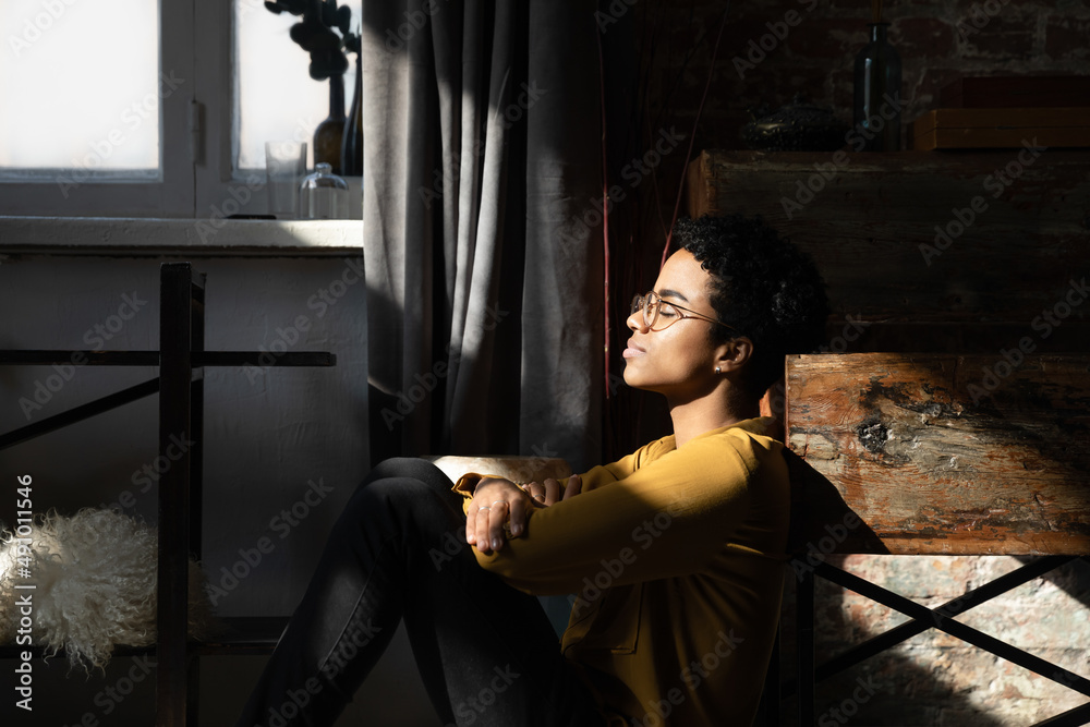 Peaceful happy young African woman, millennial girl sitting on floor in loft interior room, leaning back on shabby wooden furniture, relaxing with closed eye with day light shining on face