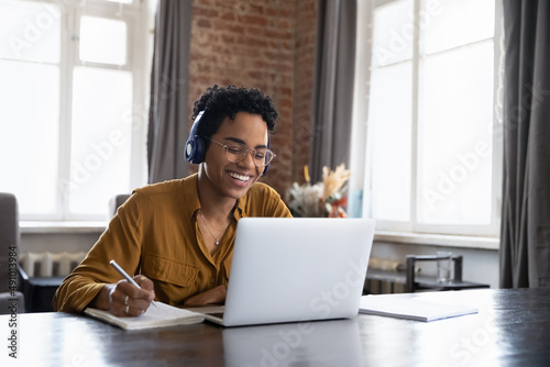 Cheerful Black college student in earphones attending virtual class, writing notes, watching webinar on Internet, studying online. African worker in headphones talking to customer on video call photo