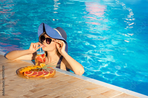 Young girl in a blue hat relaxes in the pool and drinks a cool cocktail of fresh fruit.
