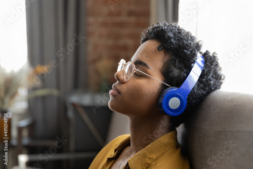 Peaceful relaxed African girl in wireless headphones sleeping to lounge music songs, resting on sofa at home. Calm sleepy fan listening to chill out tunes with closed eyes for relaxation