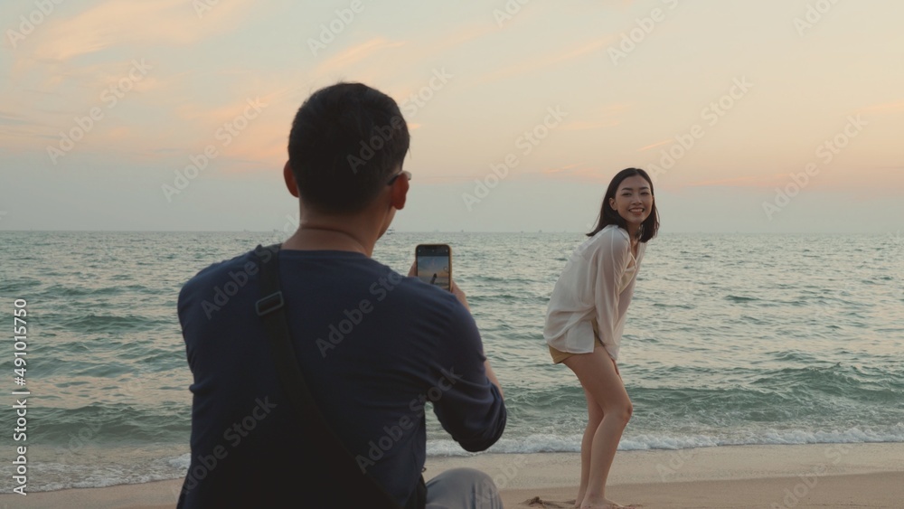 Happy man using smartphone camera making photo to woman at the beach and having fun together on beach in holiday vacation time at sunset, Young Asian woman and man in summer travel outdoor Happy man using smartphone camera making photo to woman at the beach and having fun together on beach in holiday vacation time at sunset, Young Asian woman and man in summer travel outdoor