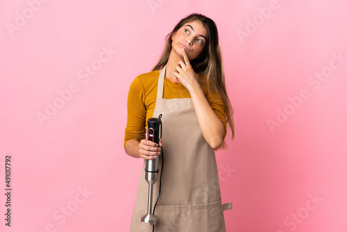Young chef woman using hand blender isolated on pink background having doubts while looking up
