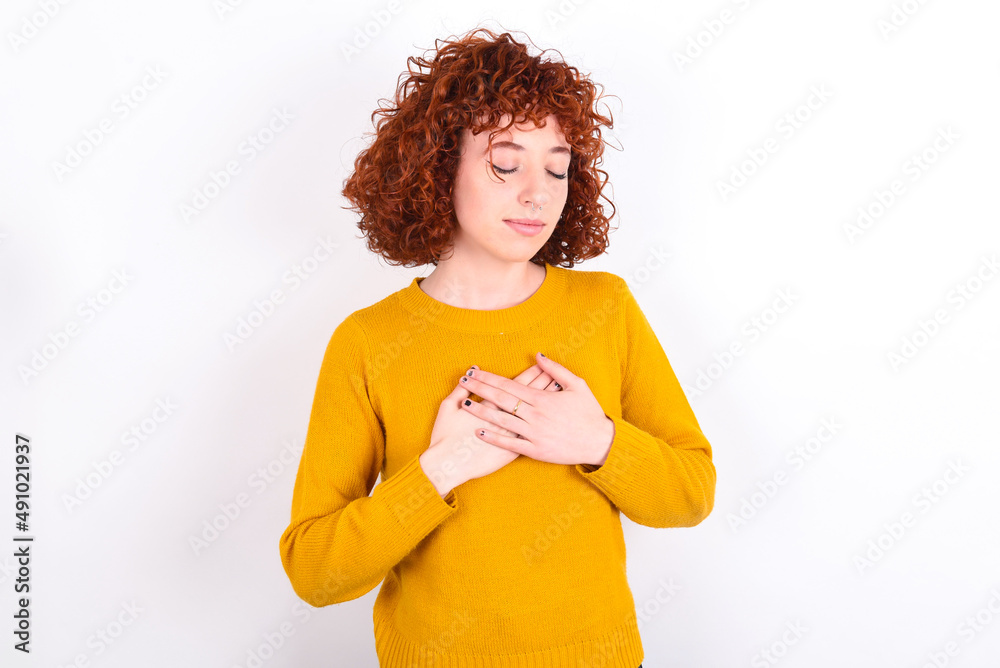 young redhead girl wearing yellow sweater over white background closes eyes and keeps hands on chest near heart, expresses sincere emotions, being kind hearted and honest. Body language 
