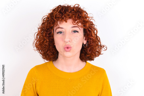 young redhead girl wearing yellow sweater over white background, keeps lips as going to kiss someone, has glad expression, grimace face. Standing indoors. Beauty concept.