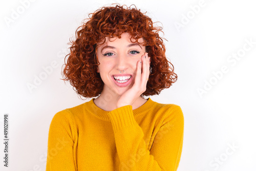 Shocked, astonished young redhead girl wearing yellow sweater over white back looking surprised in full disbelief wide open mouth with hand near face. Positive emotion facial expression body language.