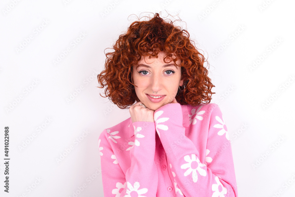 Satisfied young redhead girl wearing pink floral t-shirt over white background touches chin with both hands, smiles pleasantly, rejoices good day with lover Satisfied young redhead girl wearing pink floral t-shirt over white background touches chin with both hands, smiles pleasantly, rejoices good day with lover