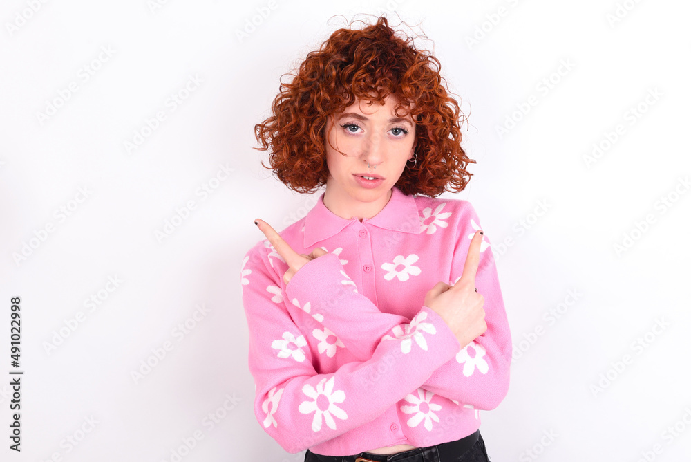 Serious young redhead girl wearing pink floral t-shirt over white background crosses hands and points at different sides hesitates between two items. Hard decision concept Serious young redhead girl wearing pink floral t-shirt over white background crosses hands and points at different sides hesitates between two items. Hard decision concept