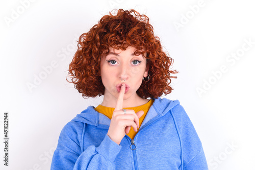 Surprised young redhead girl wearing blue jacket over white background makes silence gesture, keeps finger over lips and looks mysterious at camera photo