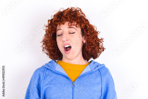 young redhead girl wearing blue jacket over white background yawns with opened mouth stands. Daily morning routine