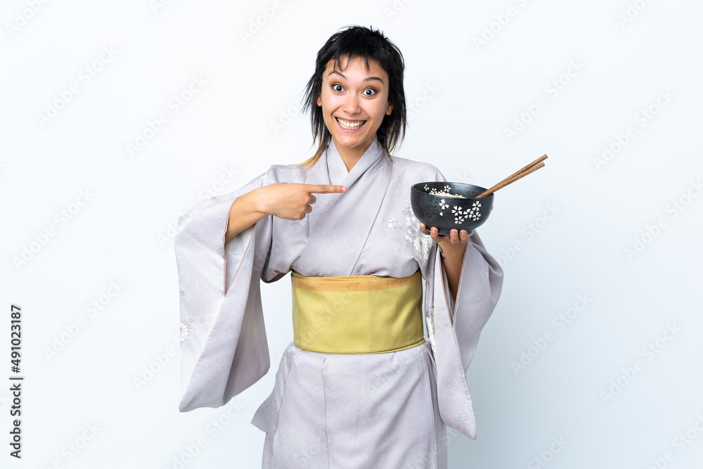 Young woman wearing kimono holding a bowl of noodles over isolated white background with surprise facial expression Young woman wearing kimono holding a bowl of noodles over isolated white background with surprise facial expression