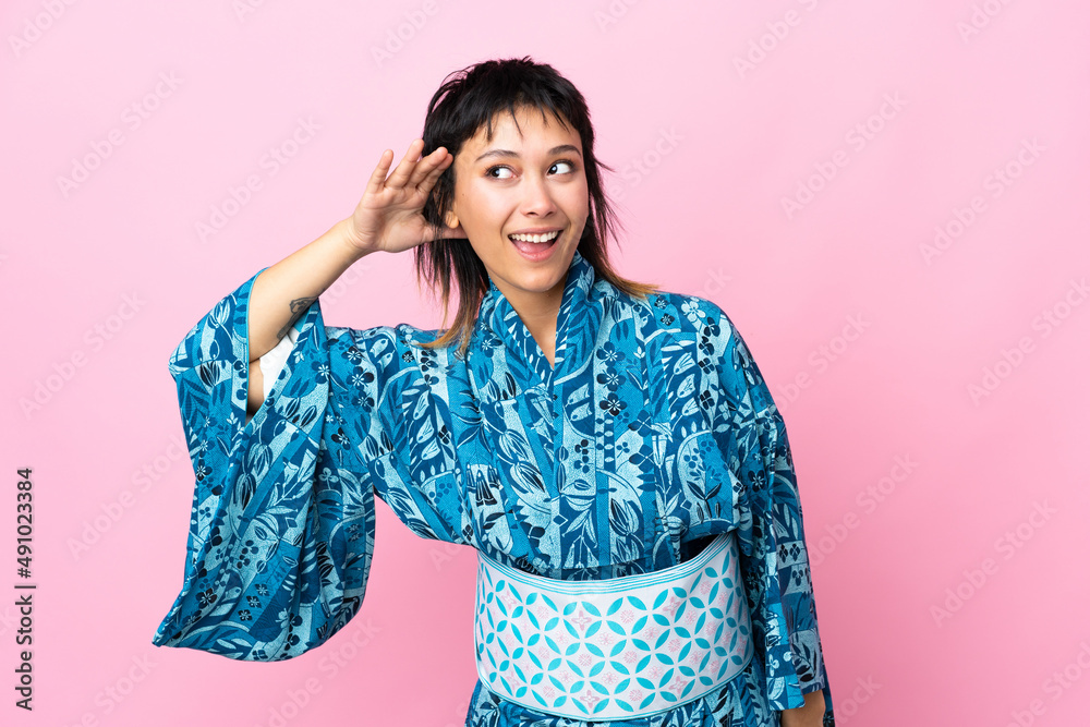 Young woman wearing kimono over isolated blue background listening to something by putting hand on the ear Young woman wearing kimono over isolated blue background listening to something by putting hand on the ear