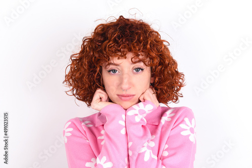Portrait of sad young redhead girl wearing pink floral t-shirt over white background hands face