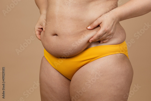 Cropped image of overweight woman, tucking, hiding fat naked big excessive belly with navel in yellow pants. Dangling down stomach, big size tummy. Drag away of abdomen. Go on diet, liposuction