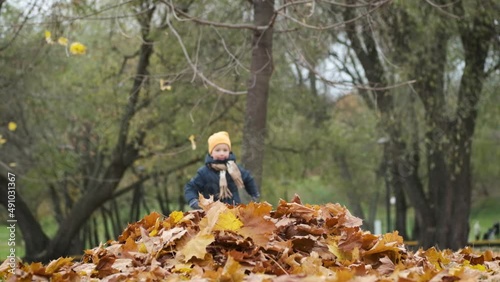 Cute child boy running and jumping into pile of autumn leaves in city park. Joys of fall season in nature outdoor. Slow motion.