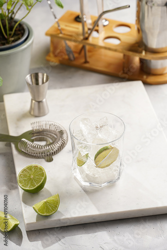 A glass with ice cubes  lime and refreshing beverage on marble board  shaker and bartender equipment