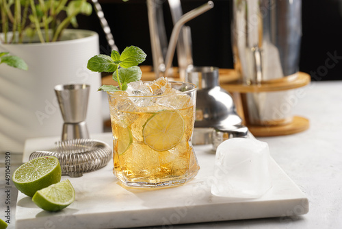 A glass with ice cubes, lime and refreshing orange beverage with mint on marble board, shaker and bartender equipment