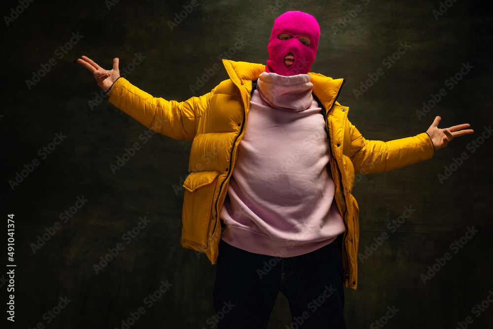 Φωτογραφία Studio shot of young anonymous man wearing pink balaclava and yellow down jacket, coat isolated on dark vintage background