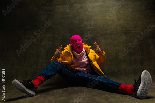 Studio shot of young anonymous man wearing pink balaclava and yellow down jacket, coat isolated on dark vintage background. Concept of safety, art, fashion photo
