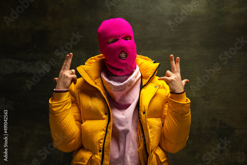 Studio shot of young anonymous man wearing pink balaclava and yellow down jacket, coat isolated on dark vintage background. Concept of safety, art, fashion photo