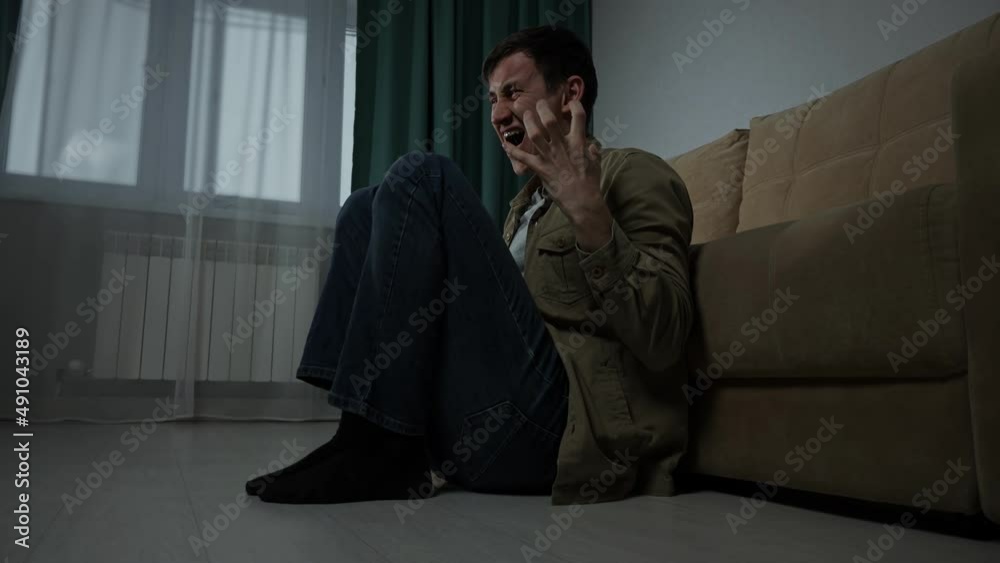 Depressed sad man with hugging the knees sits on floor near sofa in living room. Tired lost guy being alone. Depressed sad man with hugging the knees sits on floor near sofa in living room. Tired lost guy being alone.